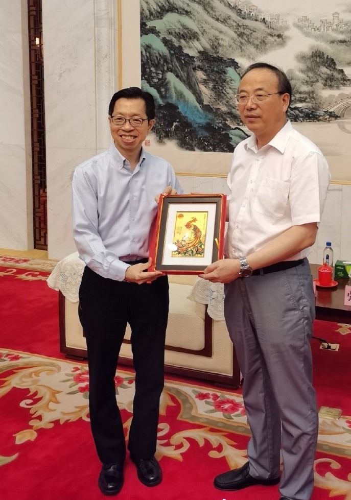 Two men holding a framed golden artwork in a room with a red patterned carpet.